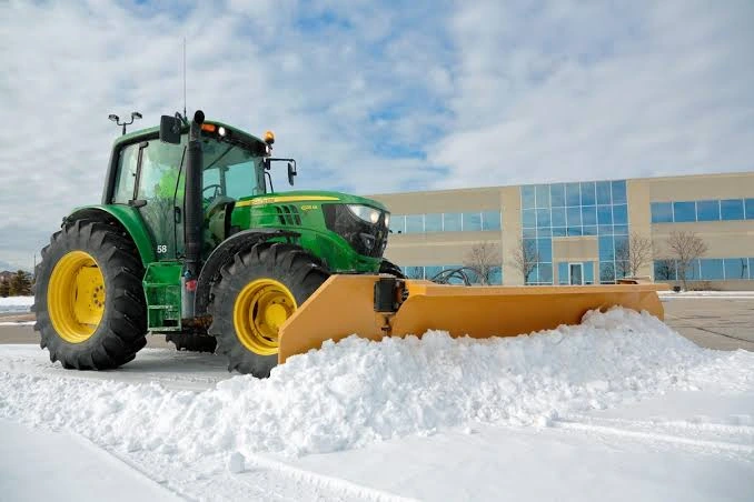 A green tractor fitted with a wide yellow snowplow clearing snow beside a commercial building.